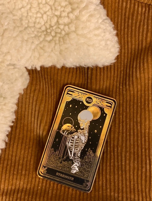 hard enamel pin of strength tarot card from the Marigold Tarot deck by Amrit Brar and 13th Press. Black pin with gold and white ink. Pin for jackets and bags