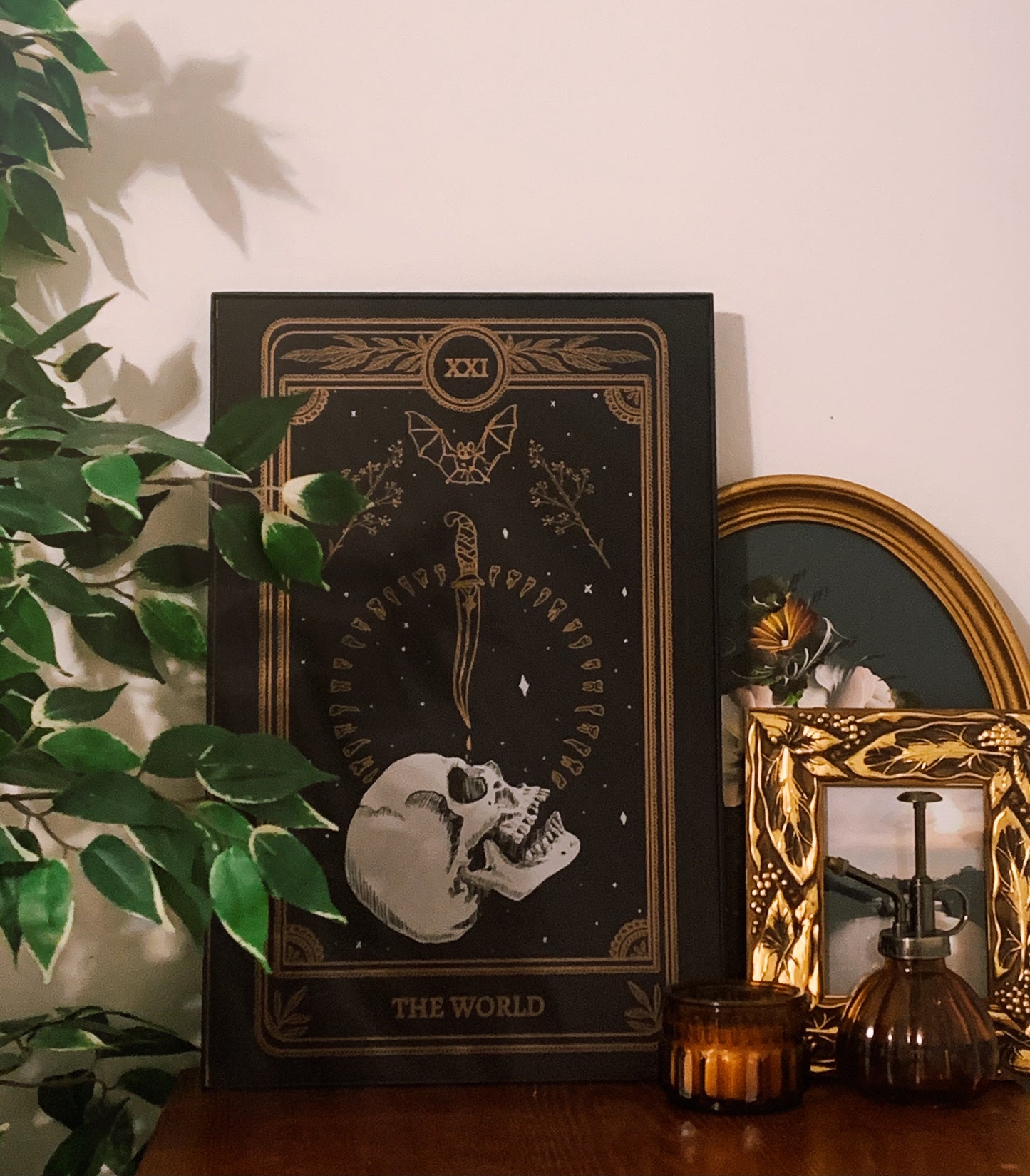 tarot card art print of the World tarot card from the Marigold Tarot deck by Amrit Brar and 13th Press. Candle light and eucalyptus foliage in the background. Black paper with white and bronze gold ink. Skull design with knife and bat.
