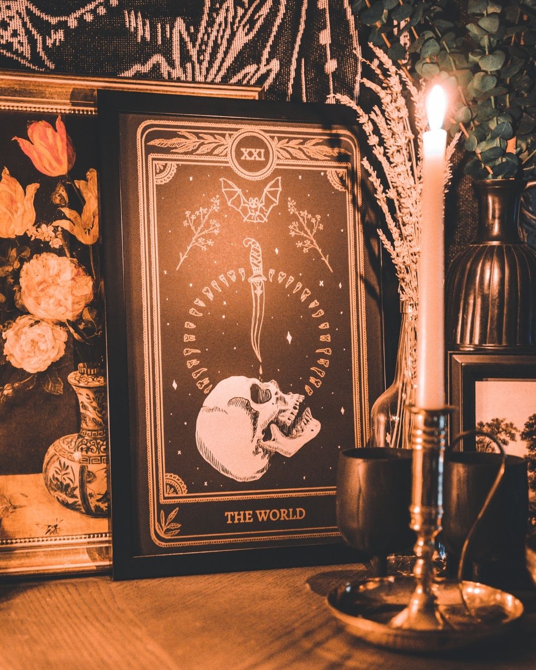 tarot card art print of the World tarot card from the Marigold Tarot deck by Amrit Brar and 13th Press. Candle light and eucalyptus foliage in the background. Black paper with white and bronze gold ink. Skull design with knife and bat.