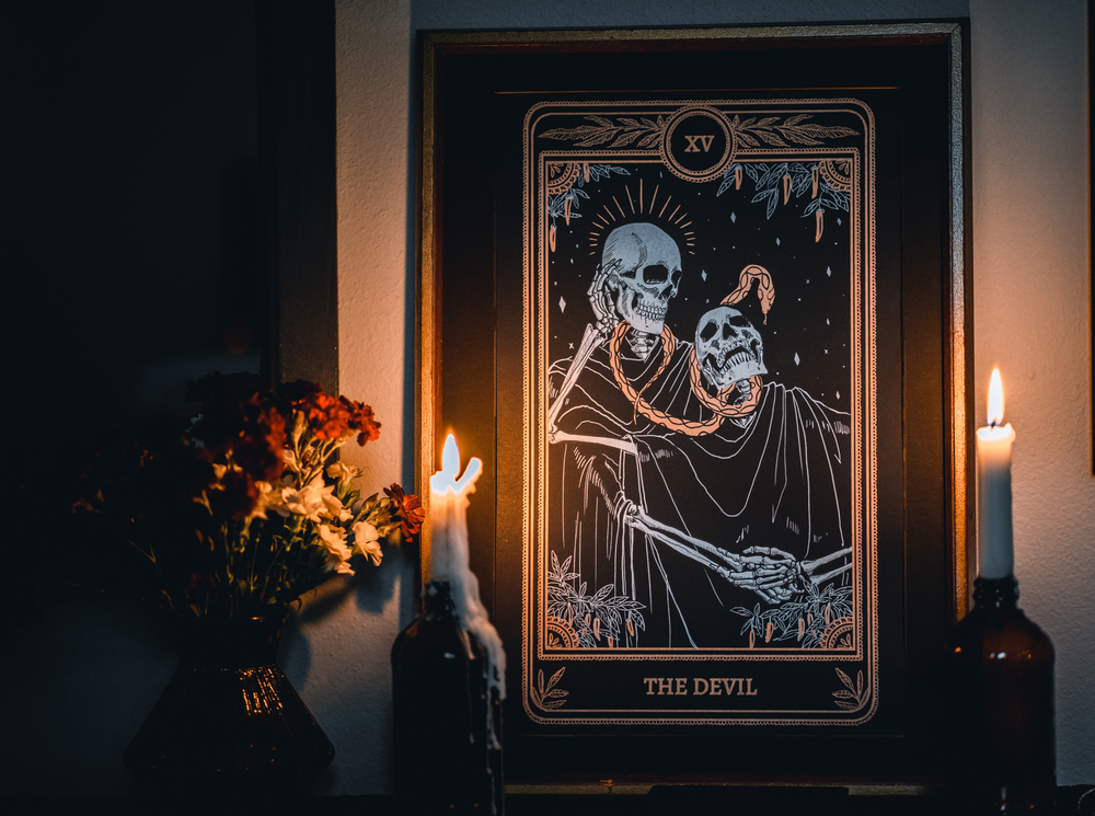 devil tarot card design from the Marigold Tarot deck by Amrit Brar and 13th Press. Black print with white and bronze gold ink. Candle light in background.