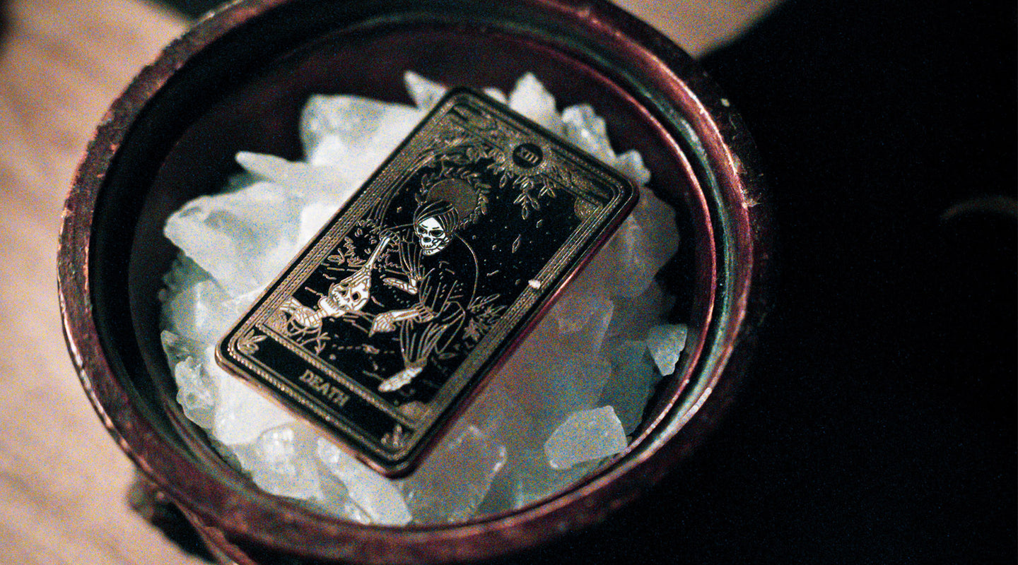 tarot death card design on black and gold metal pin. Skeleton figure holding knife above skeleton figure. Marigold Tarot Death card design.