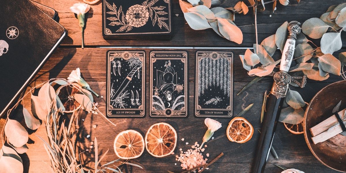 tarot card spread of Marigold Tarot deck by Amrit Brar and 13th Press. Dagger, journal, foliage, and candles in background. Black cards with gold and white ink.