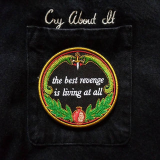 machine embroidered iron-on patch designed by Amrit Brar and 13th Press reads "the best revenge is living at all" with two serpents and pomegranate with leaves. Patch ironed onto black denim jacket pocket.