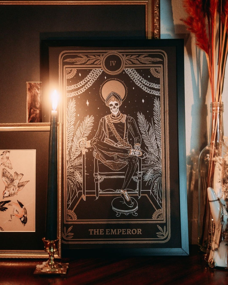 Emperor print with artwork from the Marigold Tarot deck by Amrit Brar and 13th Press on altar stand with foliage and candle light. Black print with white and gold ink of skeleton figure on throne.