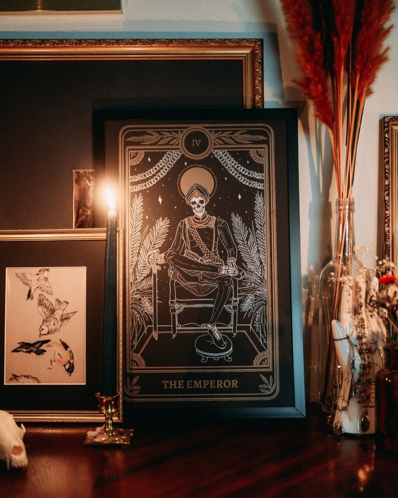 Emperor print with artwork from the Marigold Tarot deck by Amrit Brar and 13th Press on altar stand with foliage and candle light. Black print with white and gold ink of skeleton figure on throne.
