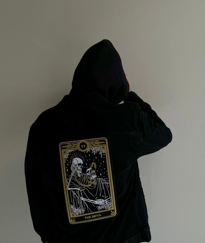 the devil tarot embroidered patch with black, gold, and white thread design from the Marigold Tarot deck by Amrit Brar and 13th Press. Iron-on patch on black denim jacket