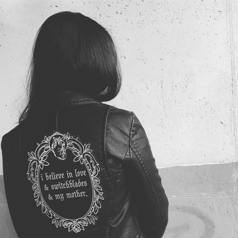 black and white iron-on embroidered back patch design from the Marigold Tarot deck by Amrit Brar and 13th Press. Text reads "i believe in love, switchblades, and my mother". Patch on black leather jacket.