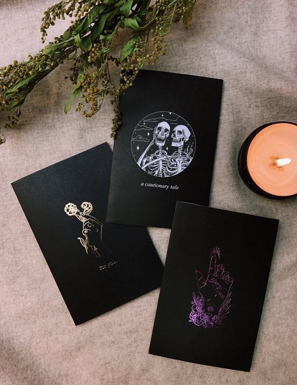 Bundle of three greeting cards, foil printed on black paper, blank inside, next to candle and green vines