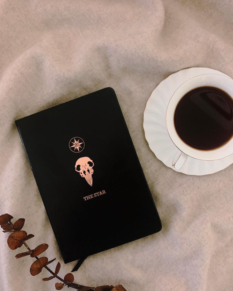 black leather journal with The Star rose gold foil designed from the Marigold Tarot deck. Bullet journal on table with coffee and foliage