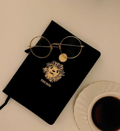 black leather journal with The Sun gold foil designed from the Marigold Tarot deck. Blank journal on table with coffee and glasses