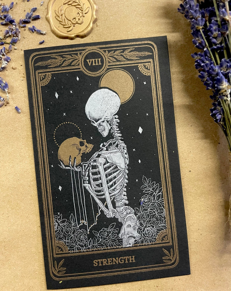 Greeting card with metallic ink printed design on black paper of the Strength tarot card from the Marigold Tarot deck by Amrit Brar and 13th press next to lavender and wax seal with skull and leaves