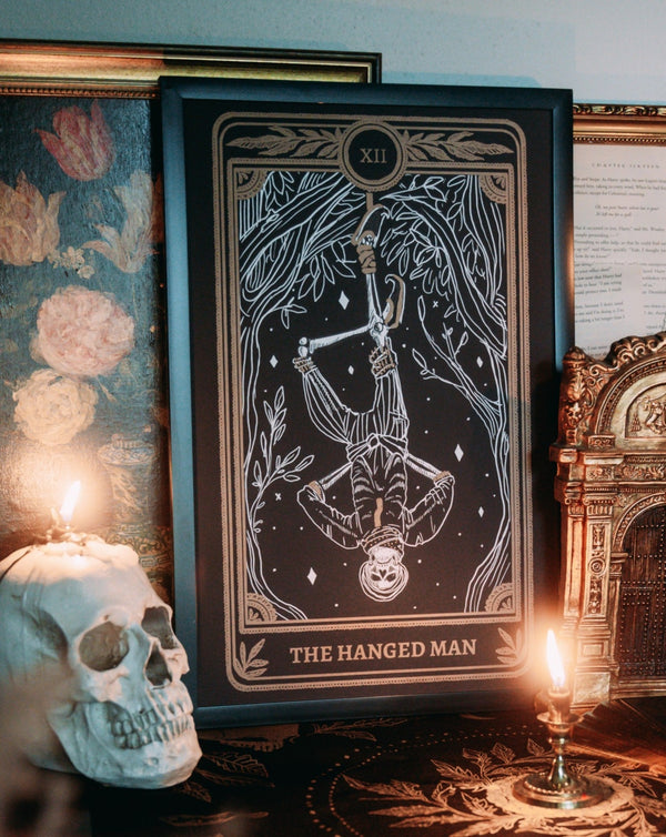 tarot art print of the Hanged Man card from the Marigold Tarot deck by Amrit Brar and 13th Press on altar with candle light and tarot card reading. Metallic ink on black paper