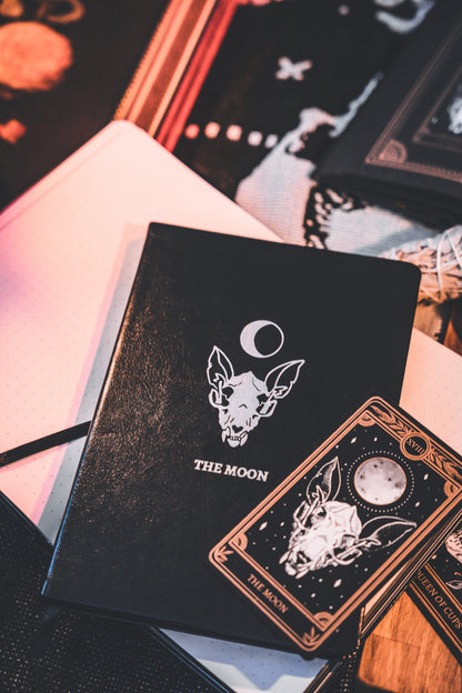 Lined journal with The Moon silver foil designed from the Marigold Tarot deck. Lined journal on altar with lit candle and The Moon tarot card from the Marigold Tarot deck