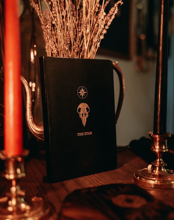 black leather journal with The Star rose gold foil designed from the Marigold Tarot deck. Bullet journal next to Marigold Tarot deck and candle light