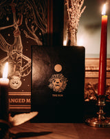 black leather journal with The Sun gold foil designed from the Marigold Tarot deck. Blank journal next to lit candle and The Hanged Man tarot print by Amrit Brar and 13th Press