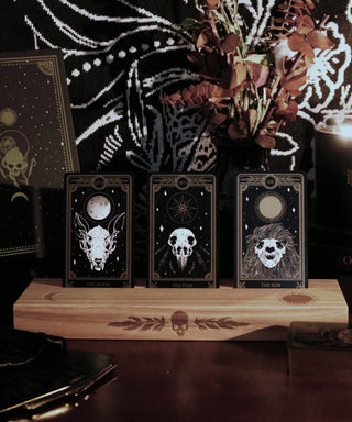 Ash wood stand featuring a hot-stamp of a skull and marigold leaves, waning crescent moon, and the sun. Excellent for holding and displaying readings while working, or for decorating your altar or desk. Comfortably fits three standard size 2.75” x 4.75” tarot or oracle cards. Stand colour may vary slightly due to differences in wood colouration. Dimensions: Approx 11.75" L x 3.5" W x 1.25" H / 30cm L x 9cm W x 3.18cm H. Composition: Ash wood. Tarot, altar, altar stand, tarot card stand, altar decor