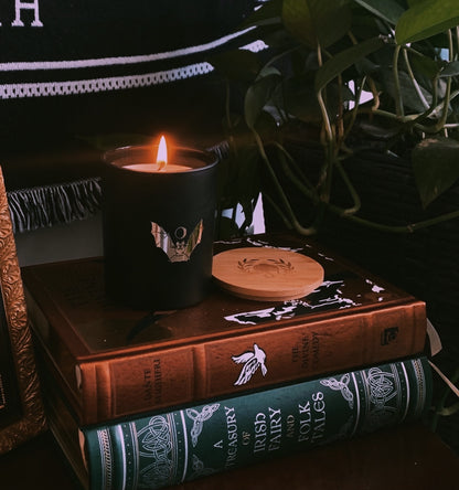 soy wax candle with matte black base and gold foil design of bat and crescent moon. T'erre de hermes scent. Displayed on altar with books and plants.