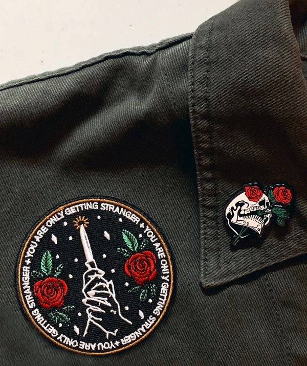 machine embroidered iron-on patch with knife and roses, text reads "you are only getting stranger" on green denim jacket with hard enamel pin of skeleton and rose.