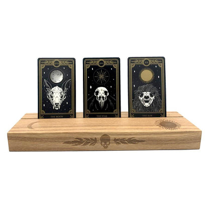 Ash wood stand featuring a hot-stamp of a skull and marigold leaves, waning crescent moon, and the sun. Excellent for holding and displaying readings while working, or for decorating your altar or desk. Comfortably fits three standard size 2.75” x 4.75” tarot or oracle cards. Stand colour may vary slightly due to differences in wood colouration. Dimensions: Approx 11.75" L x 3.5" W x 1.25" H / 30cm L x 9cm W x 3.18cm H. Composition: Ash wood. Tarot, altar, altar stand, tarot card stand, altar decor