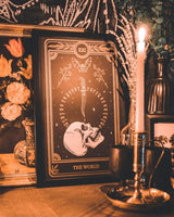 tarot card art print of the World tarot card from the Marigold Tarot deck by Amrit Brar and 13th Press. Candle light and eucalyptus foliage in the background. Black paper with white and bronze gold ink. Skull design with knife and bat.