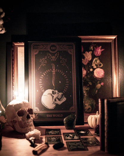tarot card art print of the World tarot card from the Marigold Tarot deck by Amrit Brar and 13th Press. Candle light and eucalyptus foliage in the background. Black paper with white and bronze gold ink. Skull design with knife and bat.