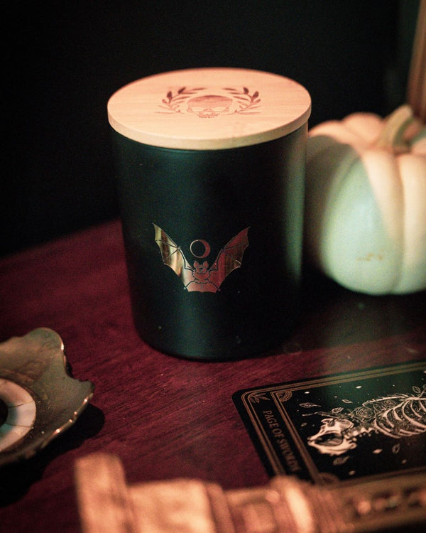 soy wax candle with matte black base and gold foil design of bat and crescent moon. T'erre de hermes scent. Pumpkin and tarot cards in background.