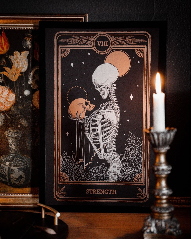 Tarot Strength card print from the Marigold Tarot deck by Amrit Brar and 13th Press with candle light on altar. Black and dim gold ink. With skeleton holding skull.