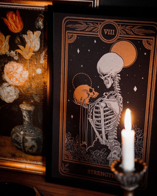 Tarot Strength card print from the Marigold Tarot deck by Amrit Brar and 13th Press with candle light on altar. Black and dim gold ink. With skeleton holding skull
