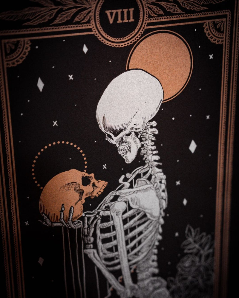 Tarot Strength card print from the Marigold Tarot deck by Amrit Brar and 13th Press with candle light on altar. Black and dim gold ink. With skeleton holding skull